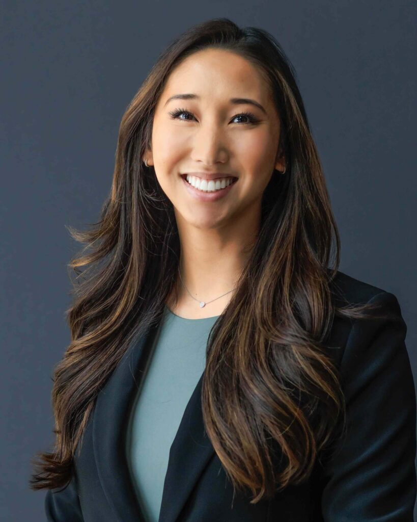 A person with long, wavy hair is smiling and wearing a black blazer over a green top against a dark background, embodying the elegance often associated with Suncadia real estate.