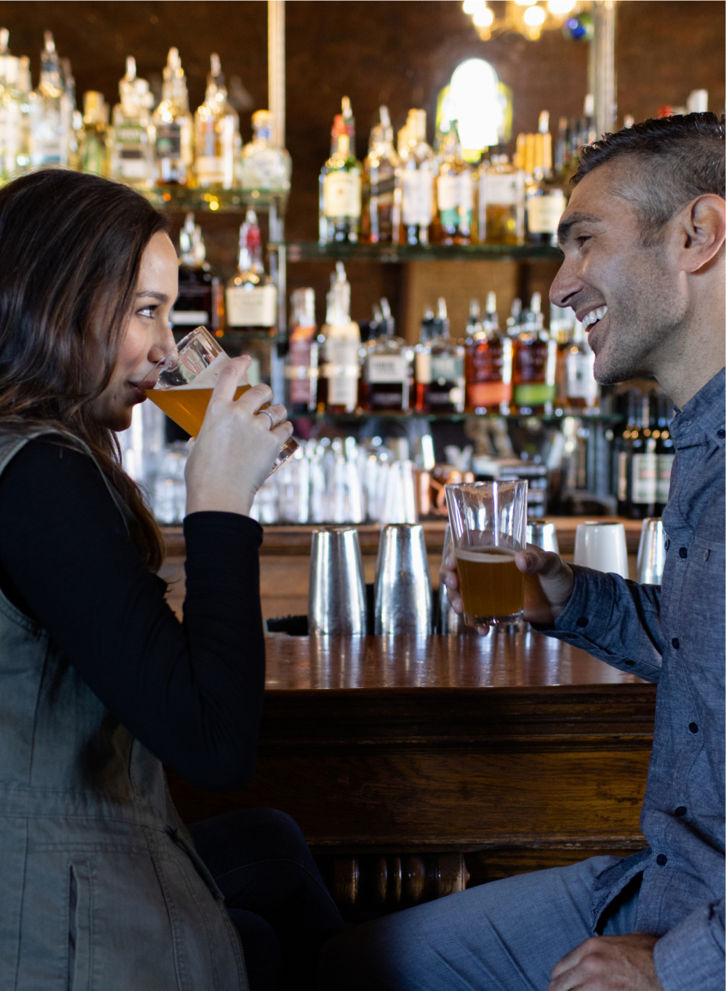 Two people sitting at a bar in a cozy Cle Elum restaurant, drinking beer and smiling at each other. Bottles and glasses glimmer softly in the background.