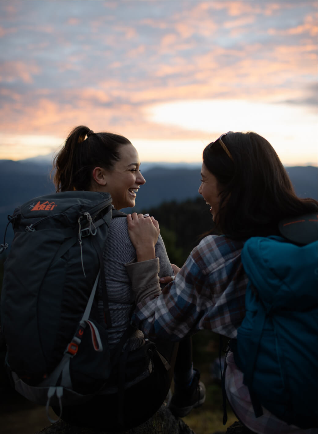 Two people with backpacks sit on a rock, smiling at each other as the sunset paints the sky. Their adventure began at Suncadia Resort, Washington, and tales of Cle Elum restaurants filled their conversations.