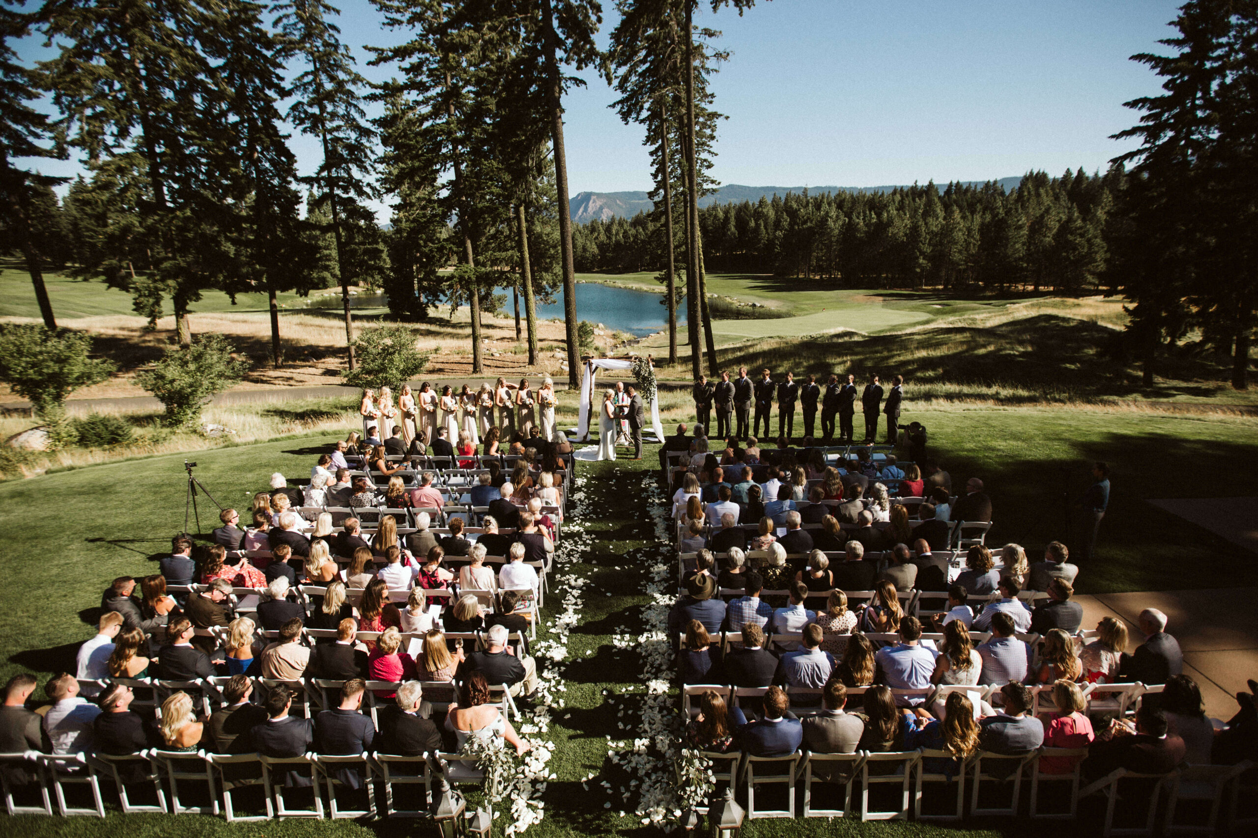 The outdoor wedding ceremony at Suncadia Resort features guests seated in rows facing a lovely arbor. The picturesque setting includes a grassy area, bordered by tall trees with a serene lake in the background, making it an unforgettable experience.