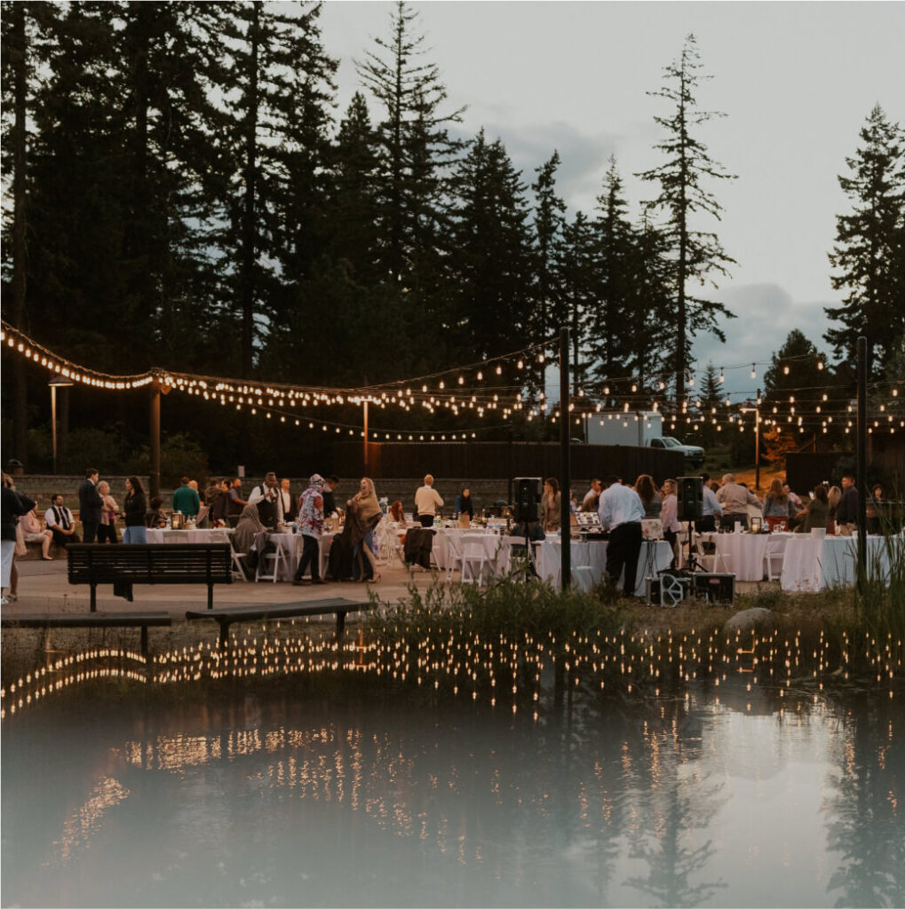 An enchanting outdoor wedding reception unfolds at dusk at Suncadia Resort, with guests mingling under twinkling string lights, surrounded by majestic trees and a serene water feature reflecting the magic of the evening.
