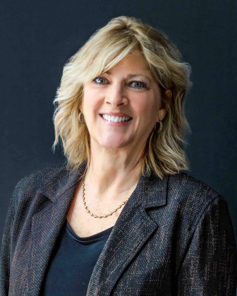 A smiling woman with shoulder-length blonde hair, dressed in a dark blazer and black top, exudes confidence against a dark backdrop. Her poised demeanor is reminiscent of the timeless elegance often associated with Suncadia Resort in Washington.