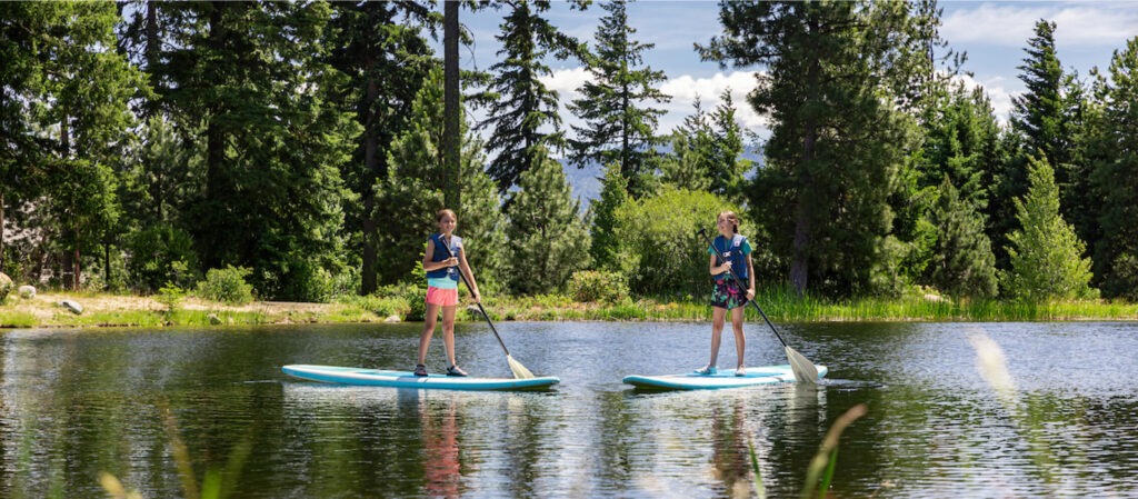 Two people are stand-up paddleboarding on a calm lake surrounded by trees under a clear sky, enjoying the serene beauty of Suncadia Resort.