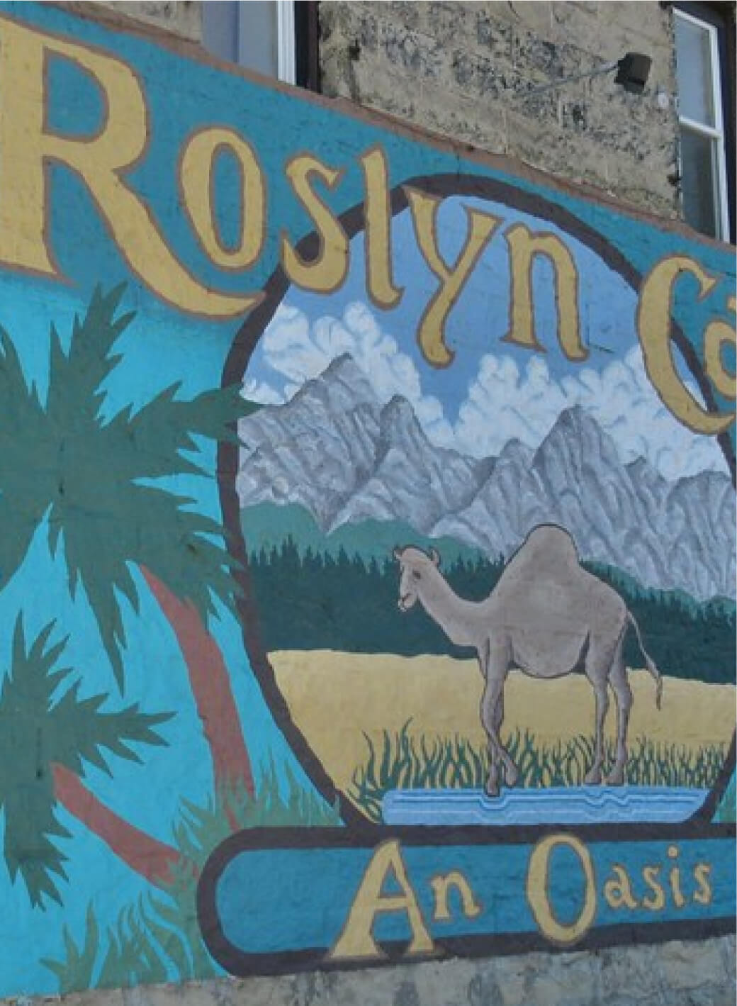 A mural on a wall showcases a camel in a desert scene with mountains, palm trees, and text reading "Roslyn Cafe An Oasis," capturing the essence of Cle Elum restaurants near the famed Suncadia Resort.