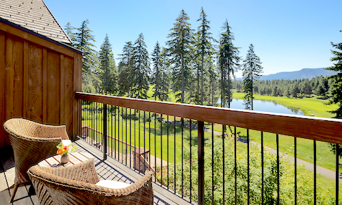 A balcony with two wicker chairs overlooks a verdant landscape at Suncadia, revealing tall trees and a serene pond under the clear blue sky.