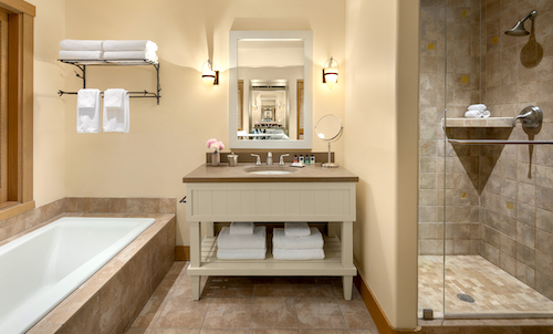 This bathroom at Suncadia Resort features a bathtub, shower, and vanity. Soft towels rest neatly on a shelf above the tub. A large mirror above the sink reflects the space, while beige tiles elegantly cover the floor and shower, adding a touch of serene luxury to your stay.