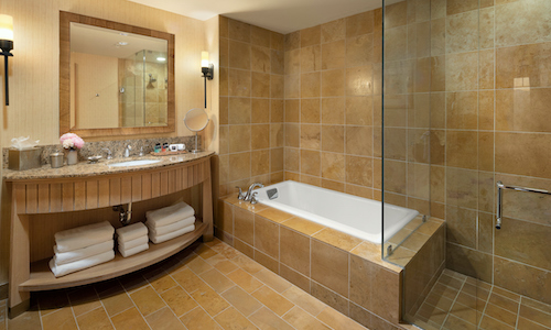 A modern bathroom, reminiscent of the luxurious design found in Suncadia Resort, features a tiled floor and walls, a bathtub with a glass screen, and a large mirror above a granite countertop. Neatly stacked towels rest on the shelf, echoing the elegance seen in Suncadia real estate.