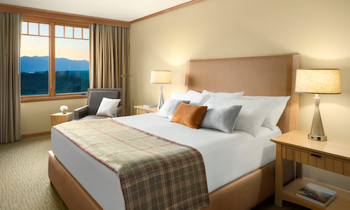 A hotel room with a large bed, plaid blanket, and plush pillows at Suncadia Resort. A window reveals a breathtaking mountain view. Lamps, a chair, and a side table complement the serene beige decor.