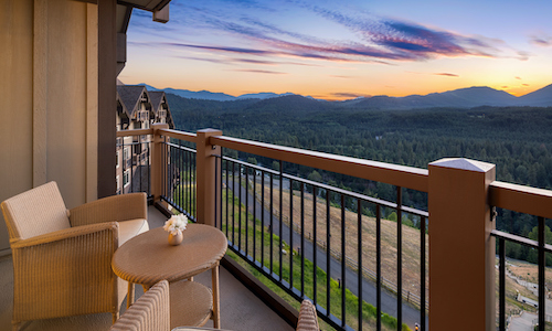 A balcony with two wicker chairs and a table overlooks a scenic landscape of mountains and forests at sunset, capturing the serene allure that defines Suncadia real estate.