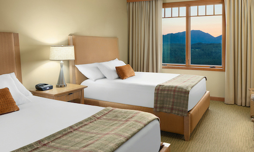 Hotel room with two double beds, a nightstand featuring a lamp and clock, and a window revealing a breathtaking mountain view at sunset. Experience the serene beauty of nature's palette at Suncadia Resort.