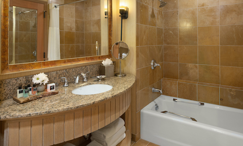 Experience a taste of Suncadia Resort luxury in this elegant hotel bathroom, featuring a sleek granite countertop, polished metal faucet, and a charming mirror surrounded by delicate flowers. Enjoy relaxing moments in the bathtub with tan tiled walls and indulge in premium toiletries.