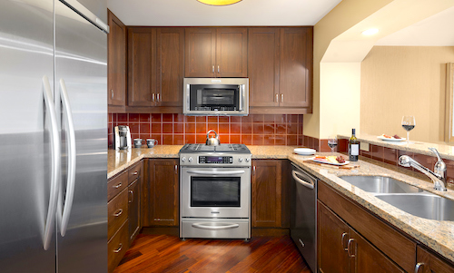 Modern kitchen with stainless steel appliances, brown cabinets, a red tile backsplash, and a double sink. Reminiscent of Suncadia Resort style, a wine glass and bottle beckon from the granite countertop.