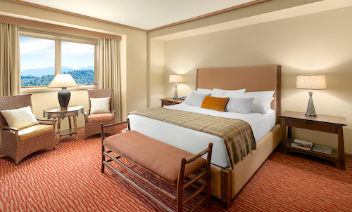 The hotel room features a large bed with decorative pillows, two chairs, and a small table, all illuminated by two lamps. A window reveals serene views of trees and hills reminiscent of Suncadia's landscapes. The red carpet with a striped pattern adds a cozy touch.