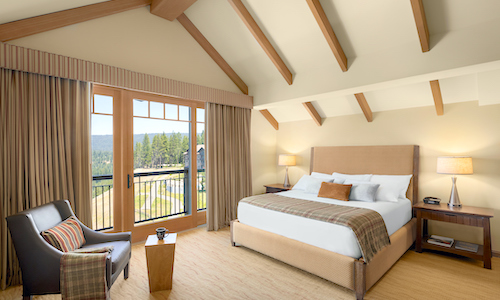 A cozy bedroom at Suncadia Resort Washington features a large bed, armchair, and wooden furniture. A glass door opens to a balcony with a scenic view of trees. Warm lighting from lamps creates an inviting atmosphere, perfect after dining at nearby Cle Elum restaurants.