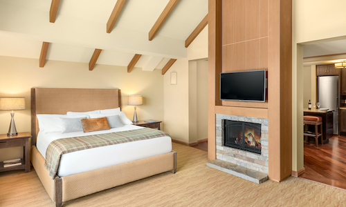 A modern bedroom at Suncadia Resort Washington features a large bed, two bedside tables, a wall-mounted TV above a fireplace, and an open view into the kitchen area with wooden flooring.