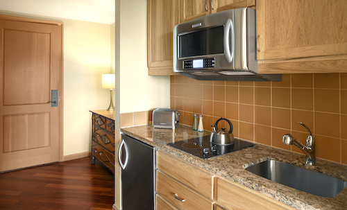 Compact kitchen with wood cabinets, granite countertop, stainless steel microwave, electric stove, sink, and a small fridge. Toaster and kettle are on the counter. Offering a taste of Suncadia real estate design with its cozy yet efficient layout. Door and lamp in the background.