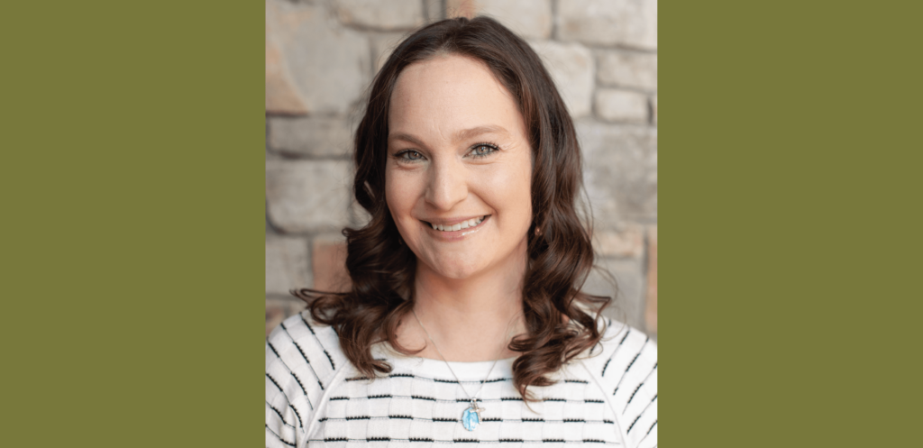 A person with wavy brown hair smiles while standing in front of a stone wall, wearing a striped shirt and a necklace, perhaps taking a break from exploring charming Cle Elum restaurants nearby.