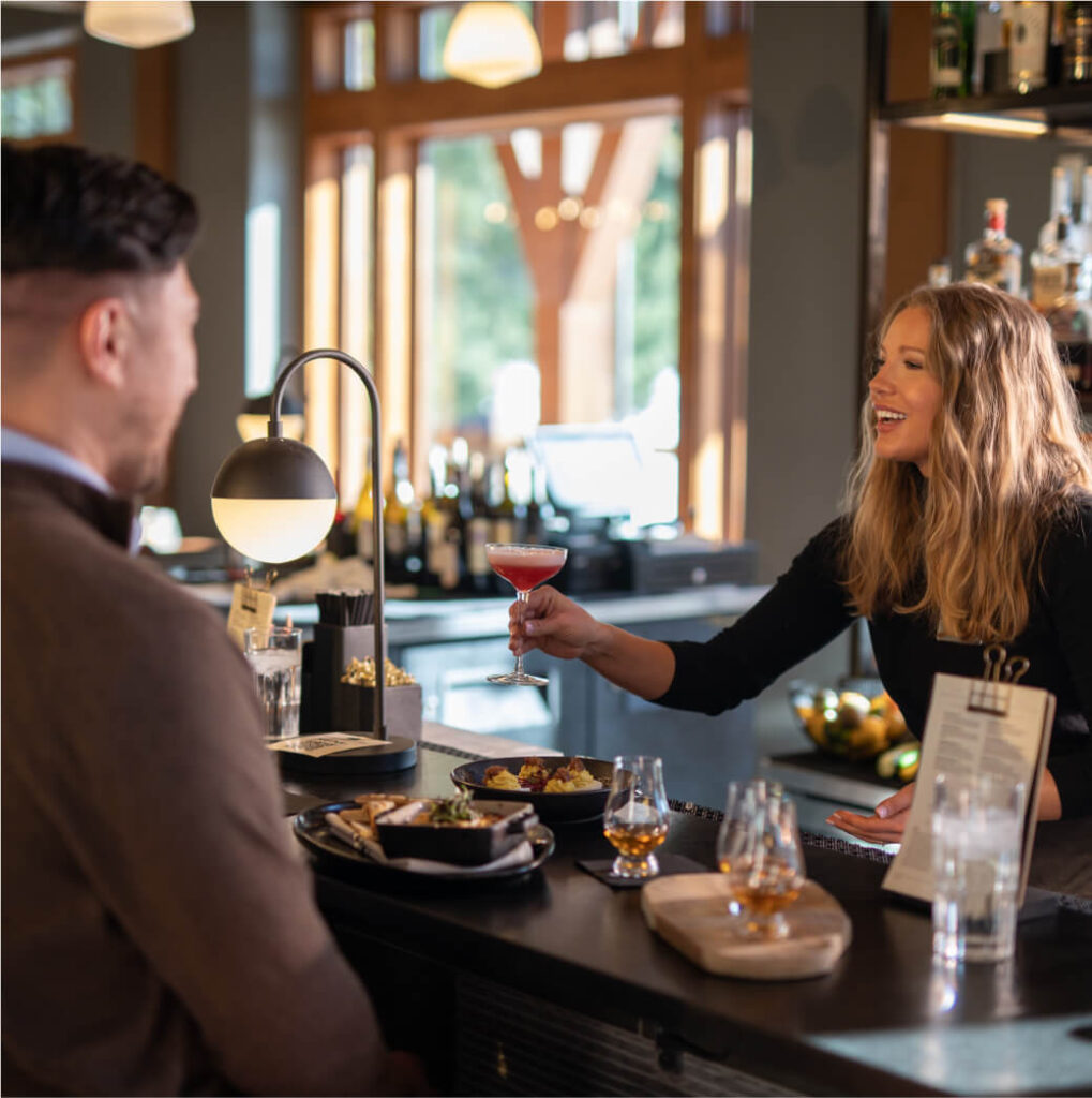 A bartender at a Cle Elum restaurant smiles as she hands a cocktail to a customer sitting at the bar. The counter is adorned with snacks and drinks, beautifully complementing the warm interior, reminiscent of the inviting ambiance found at Suncadia Resort homes.