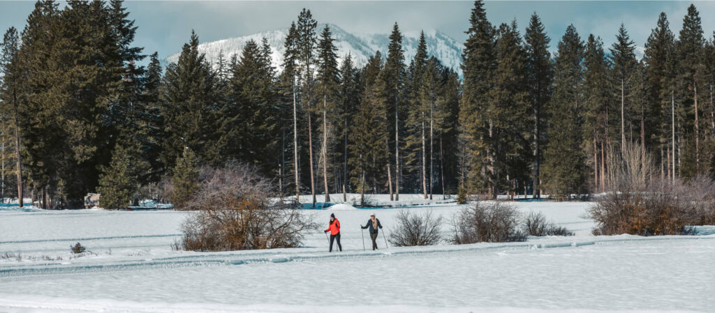 Two people stroll across a snowy landscape, framed by tall pine trees and the majestic mountain backdrop of Suncadia Resort.