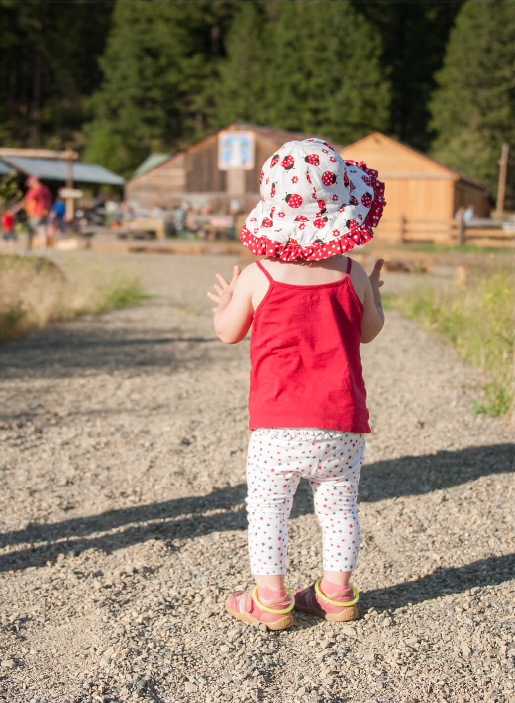 A toddler wearing a red dress, white leggings, and a floral hat walks on a gravel path in the picturesque setting of Suncadia. Wooden buildings and lush trees provide a charming backdrop, capturing the serene essence of this beautiful area.