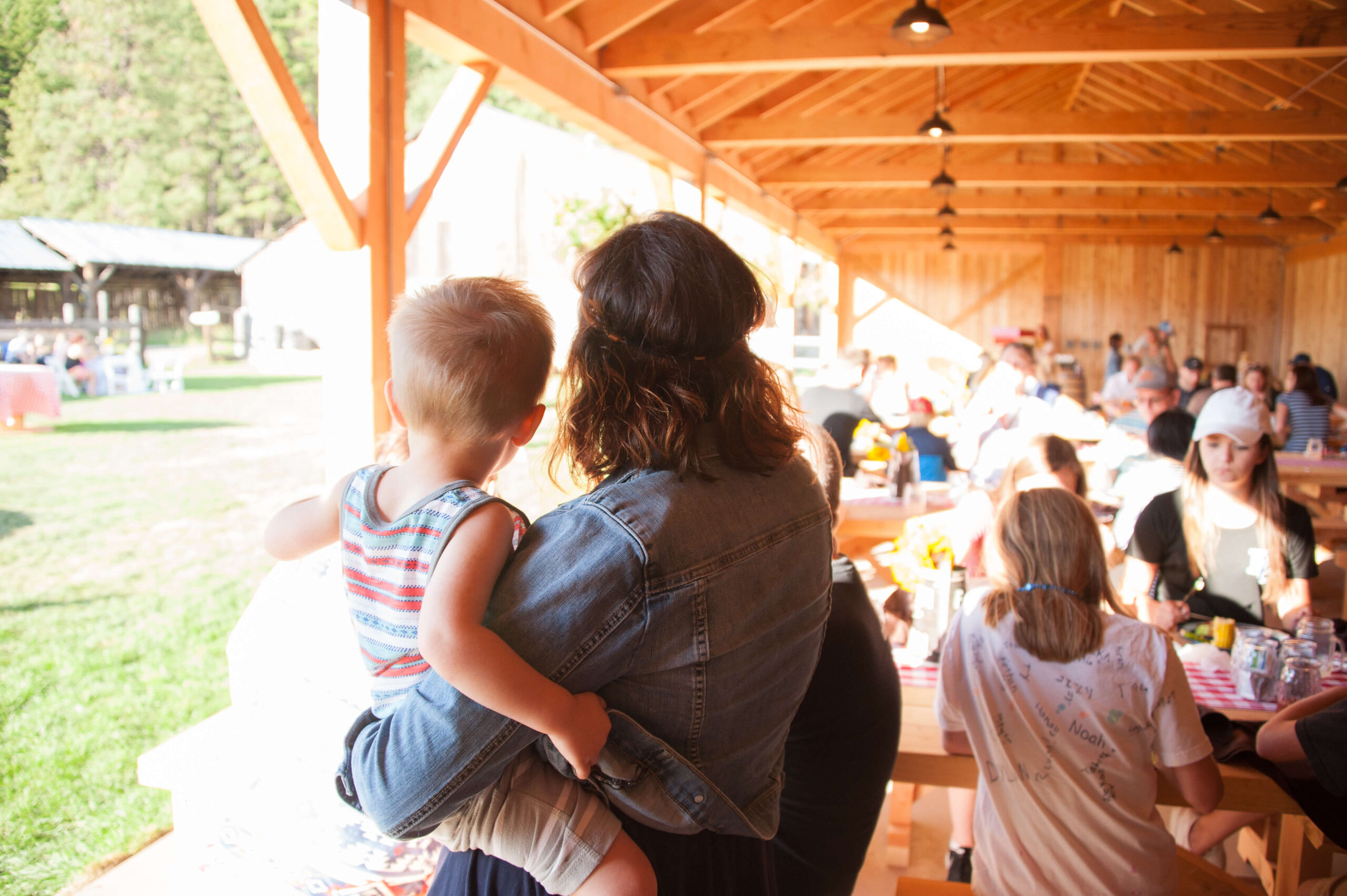 At Suncadia Resort, a woman cradles a child amid the lively buzz of a busy outdoor picnic area with wooden tables. Guests enjoy eating and socializing under the rustic charm of the wooden shelter in this vibrant slice of Washington.