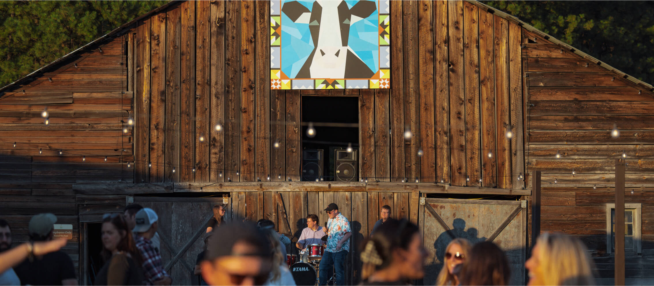 A wooden barn with a geometric animal mural at Suncadia Resort, Washington, hosts a live band performing in front. People are gathered, and string lights are hung above, creating a lively atmosphere reminiscent of the spirited gatherings at nearby Cle Elum restaurants.