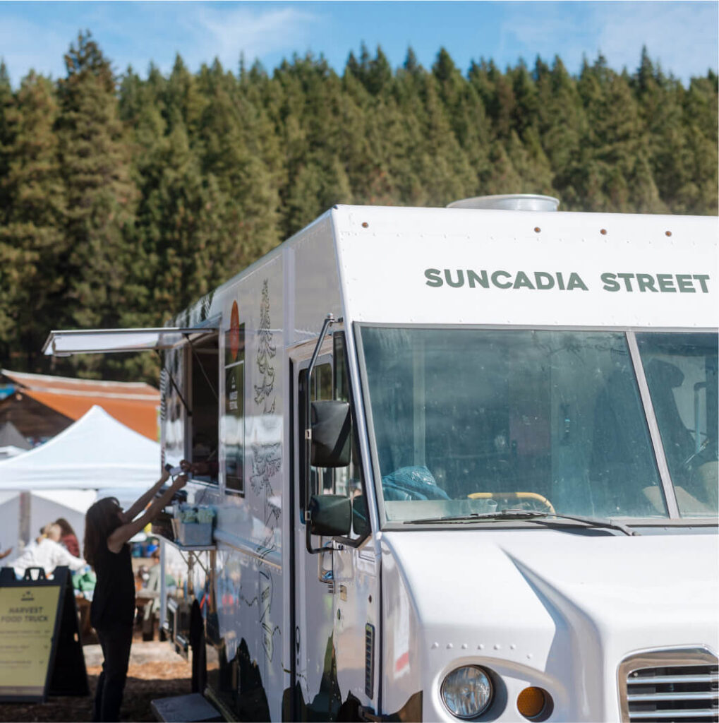 The food truck, adorned with the sign "Suncadia Street," is parked under a canopy of trees and tents. As a customer reaches up to the service window, it feels like a charming slice of Suncadia Resort life, reminiscent of Cle Elum restaurants nestled in nature's embrace.