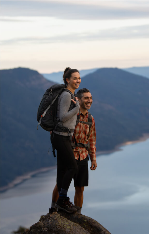 Two people with backpacks stand on a mountain cliff, overlooking a scenic view of distant mountains and a shimmering body of water near Suncadia Resort.