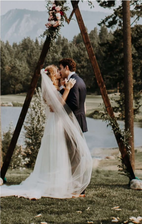 A couple embraces under a triangular wooden arch adorned with flowers at Suncadia Resort, set against a scenic outdoor backdrop of trees and a tranquil lake in Washington.