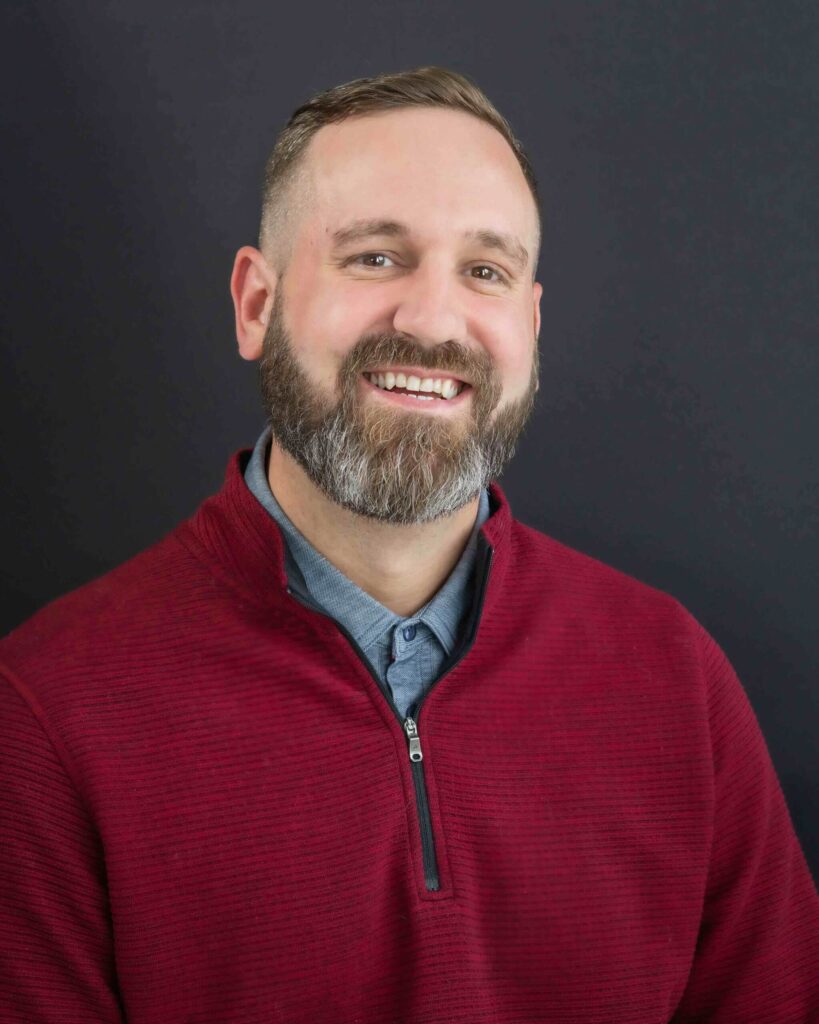 A man with a beard smiles while wearing a maroon sweater and gray shirt, reminiscent of someone enjoying the cozy ambiance of Cle Elum restaurants, all set against a dark background.