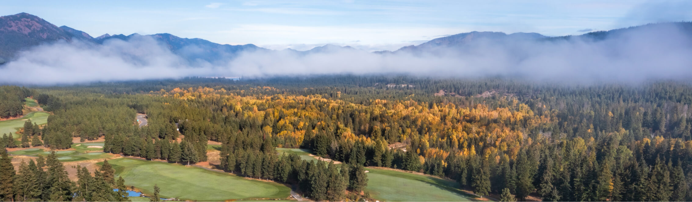 Aerial view of a forested landscape in Suncadia with colorful autumn trees, lush green fields, and distant mountains under a cloudy sky.