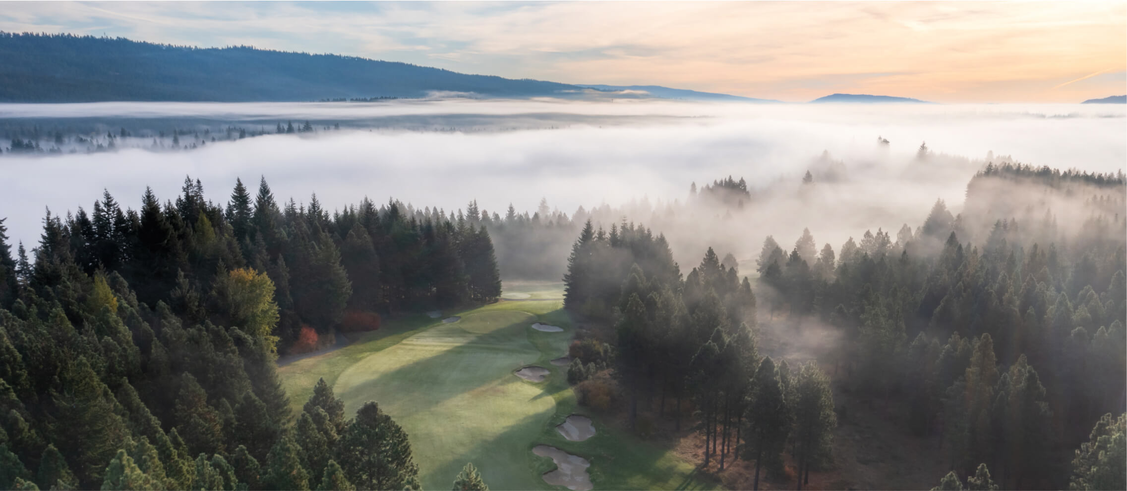 Aerial view of a golf course nestled among dense forest trees, with mist covering the landscape under a sunrise sky, captures the tranquil charm of Suncadia.