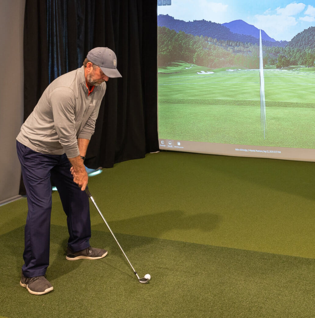 A man in golf attire prepares to swing a golf club indoors, facing a virtual golf course screen at Suncadia Resort Washington, capturing the essence of an outdoor game without leaving the comfort of this luxurious retreat.