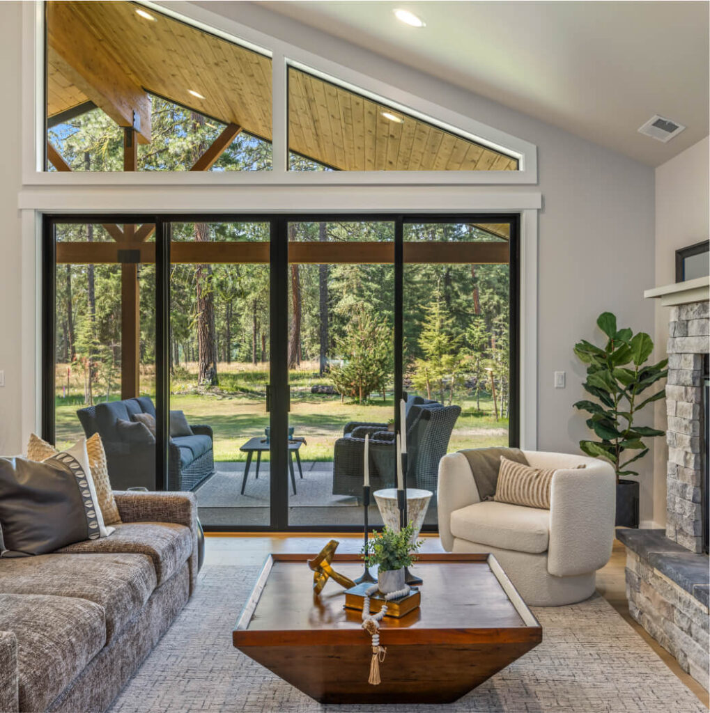 Spacious living room with large windows offering a serene forest view, perfect for enjoying the tranquility of Suncadia Resort in Washington. Features a gray sofa, cushioned chair, wooden coffee table, stone fireplace, and potted plant. Outdoor patio with furniture visible.