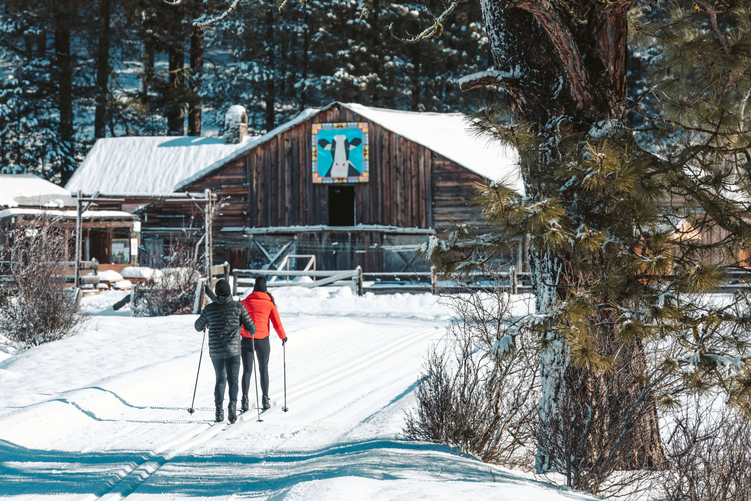 Two people in winter clothing are skiing on a snow-covered trail at Suncadia Resort, heading towards a wooden building with a colorful mural, surrounded by trees.