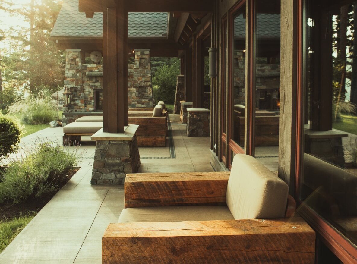 The outdoor seating area at Suncadia features wooden chairs and cushions on a covered patio. Stone pillars and lush greenery surround the space, with sunlight filtering through to create a warm atmosphere. Perfect for enjoying the serene beauty of Cle Elum's finest settings.