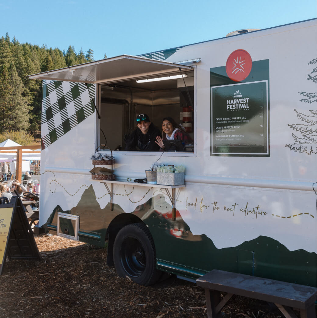 A green and white food truck at the Suncadia Harvest Festival features two people inside waving cheerfully. A sign reads "Harvest Festival," while trees from Suncadia Resort, Washington create a picturesque backdrop.