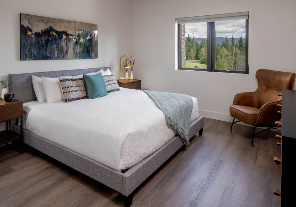 A modern bedroom at Suncadia Resort features a large bed adorned with decorative pillows and a green throw, complemented by a leather chair and a landscape painting. The window offers a serene view of trees against the backdrop of Washington's cloudy sky.