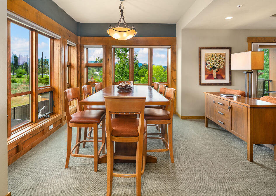 A dining room with a wooden table and chairs, large windows offering a lush view reminiscent of Cle Elum restaurants, alongside a sideboard, lamp, and wall painting that echo the elegance of Suncadia Resort.