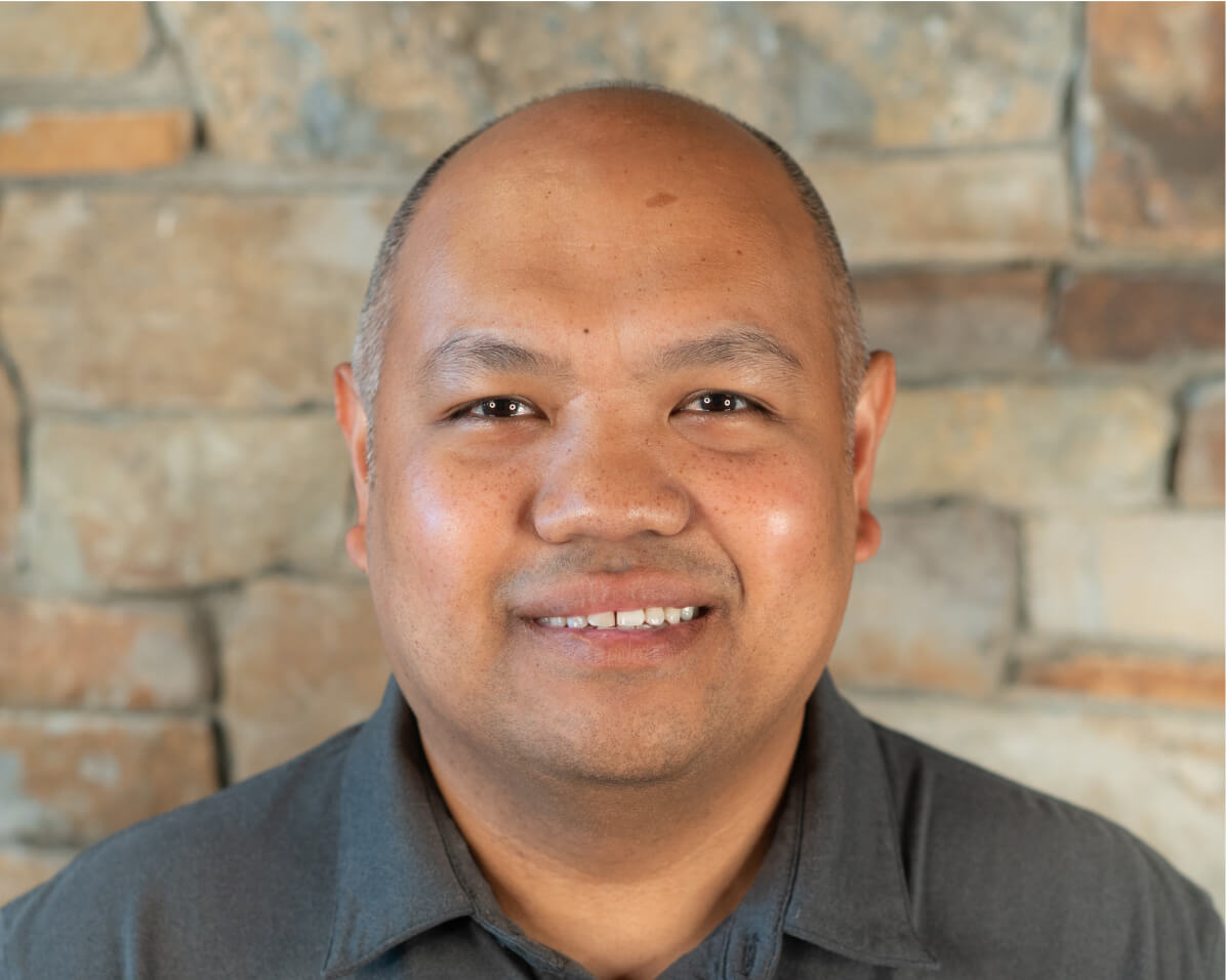 A person with a shaved head and a gray shirt smiles warmly in front of a stone wall backdrop, reminiscent of the rustic charm found at Suncadia Resort.