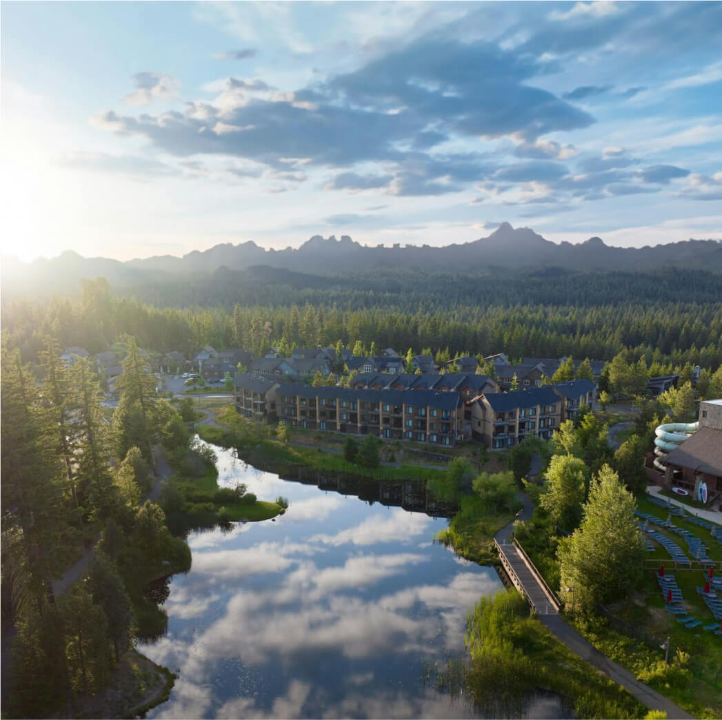 Aerial view of Suncadia Resort, nestled among trees beside a reflective lake, with mountains and a partly cloudy sky creating a stunning backdrop. Nearby, Cle Elum restaurants offer delightful dining experiences to complement your tranquil getaway.