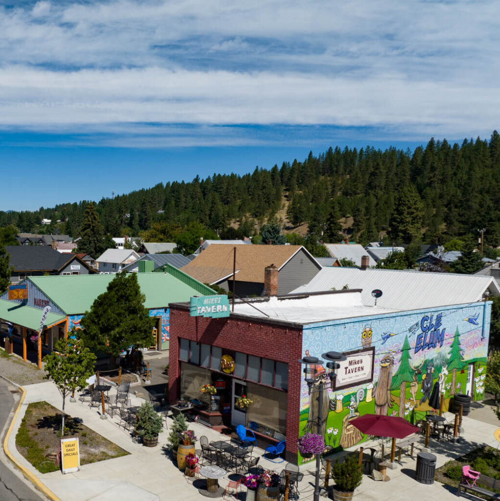 Aerial view of a small town near Suncadia Resort, showcasing a brick building adorned with colorful murals and a "Nana's Tavern" sign. Outside, tables with umbrellas create inviting spots amid lush trees and rolling hills, capturing the tranquil charm of suncadia resort washington.