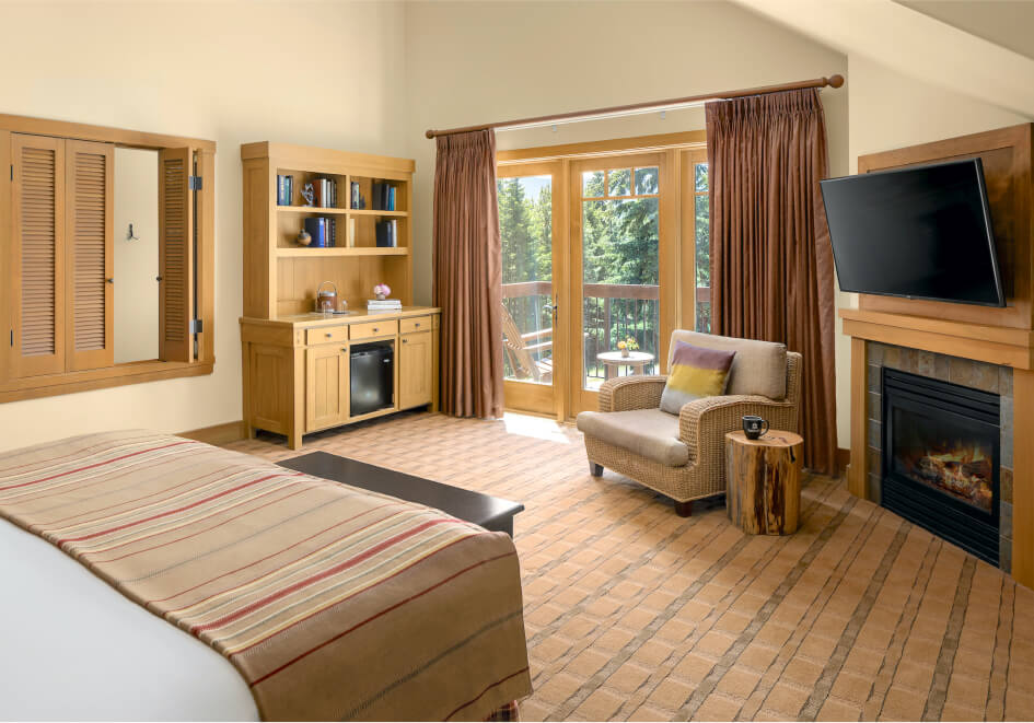 A cozy hotel room at Suncadia features a bed, wooden furniture, a fireplace, and a wall-mounted TV. Glass doors lead to a balcony overlooking lush green trees, making it an ideal retreat near Cle Elum restaurants.