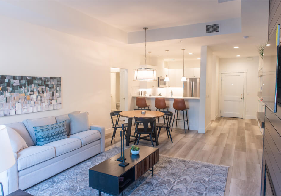 Modern living room and kitchen with a beige couch, round dining table, bar stools, and pendant lights. Contemporary decor and hardwood flooring echo the elegance found in Suncadia real estate, blending luxury with comfort in every detail.