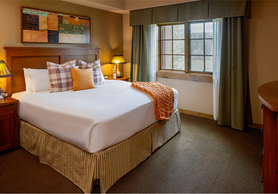 A neatly made bed with white linens, assorted pillows, and an orange throw blanket is complemented by nightstands with lamps. In the background, a window with curtains and a decorative painting completes this cozy setting at Suncadia Resort in Washington.