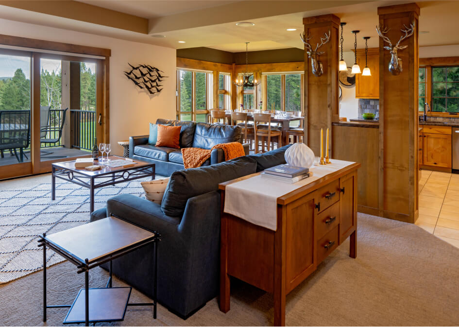 Modern open-concept living room and dining area at Suncadia Resort, featuring dark sofas, orange cushions, wooden furniture, and decorative deer head wall mounts. Enjoy the serene forest view after dining at nearby Cle Elum restaurants.