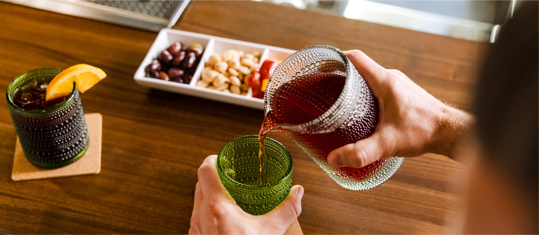 A person pours a red drink from a textured pitcher into a green glass on a wooden table at Suncadia Resort. Nearby, a drink with an orange slice and a tray of olives and nuts evoke the culinary delights familiar to Cle Elum restaurants.