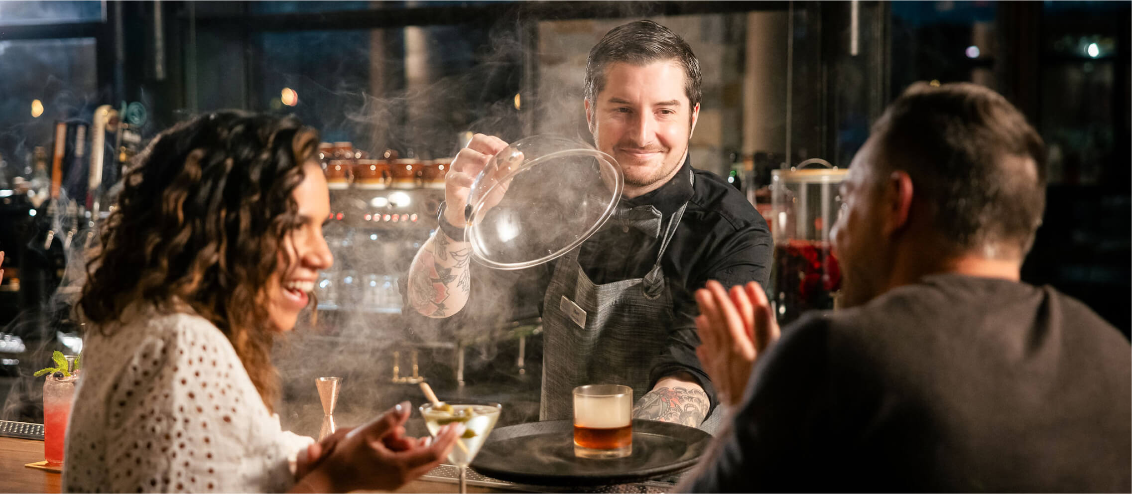 At a lively Cle Elum restaurant, a bartender serves a drink beneath a glass cloche to two patrons. The woman smiles while the man gestures animatedly, as wisps of smoke curl elegantly around the cocktail—a signature experience at Suncadia Resort.