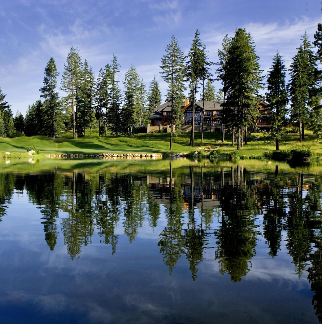 A tranquil lake reflects tall trees and a large Suncadia home under a blue sky.