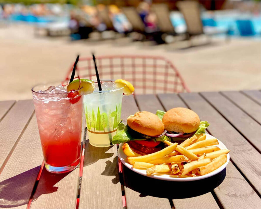 Plate with two burgers and fries on a table, paired with refreshing drinks featuring straws and lemon wedges. In the background, the inviting pool area beckons with loungers—a perfect day at Suncadia Resort Washington.